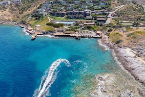 An aerial view of a coastal resort with multiple buildings, a pier extending into the sea, and a clear blue ocean.