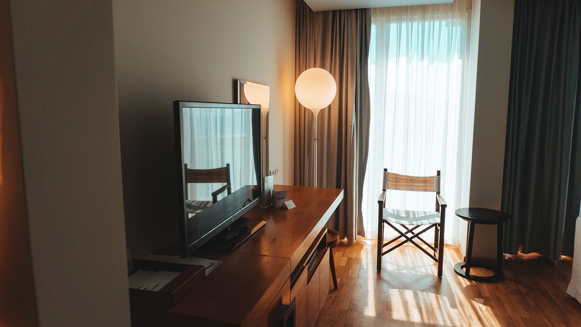 Habitación de hotel con escritorio de madera, televisor, una silla y un amplio ventanal con cortinas.