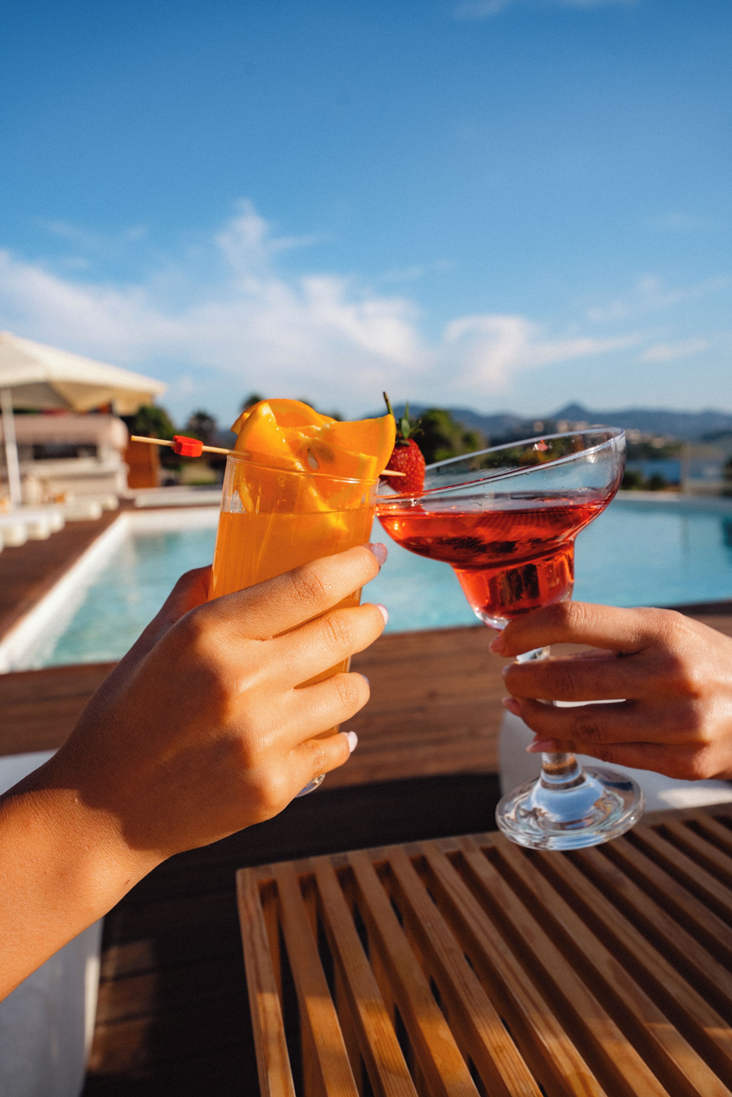 Two hands holding cocktail glasses with fruit garnishes, a pool, and a clear sky in the background.