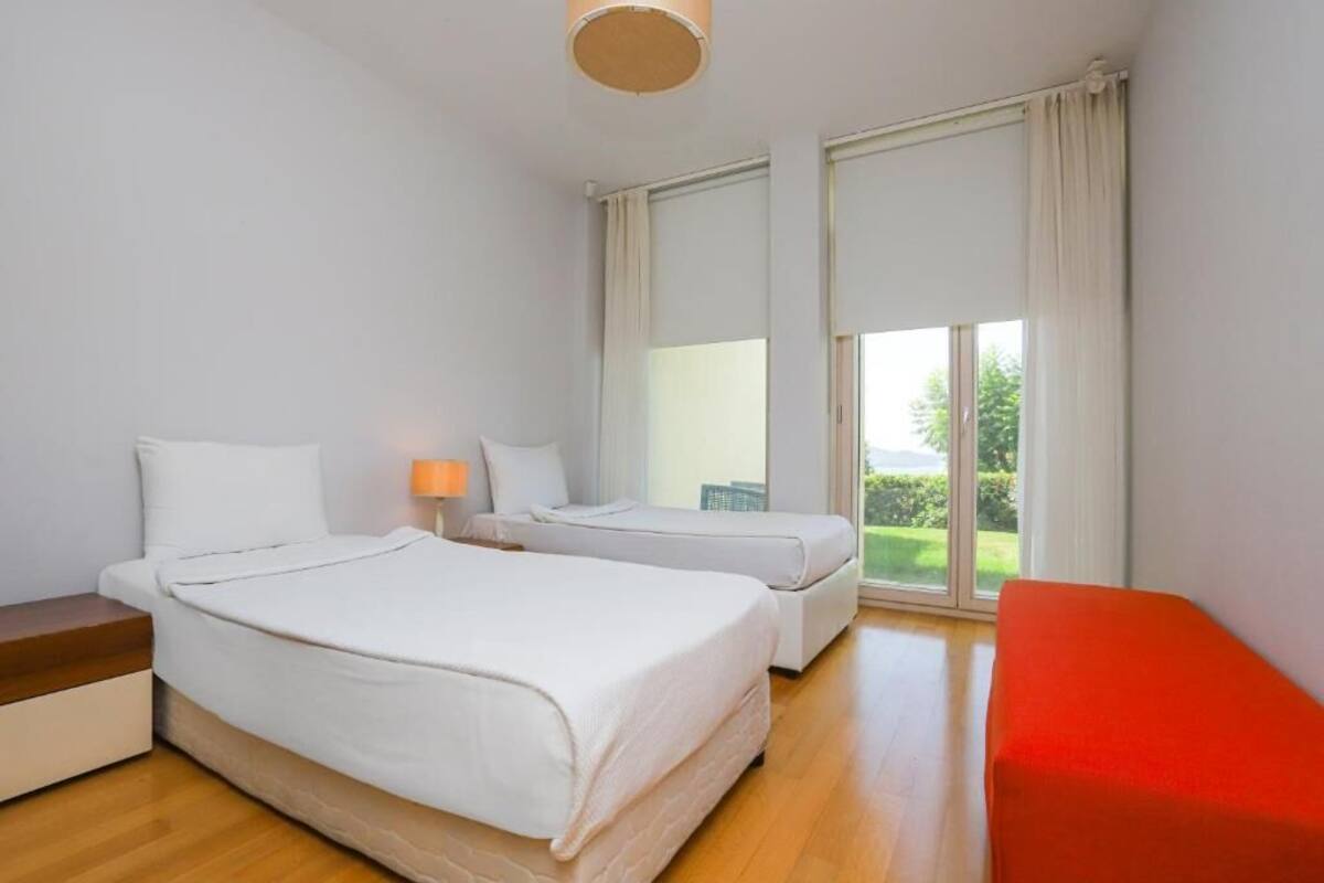 Habitación de hotel con dos camas, un banco rojo, una mesita de noche y un ventanal con vistas al verdor.