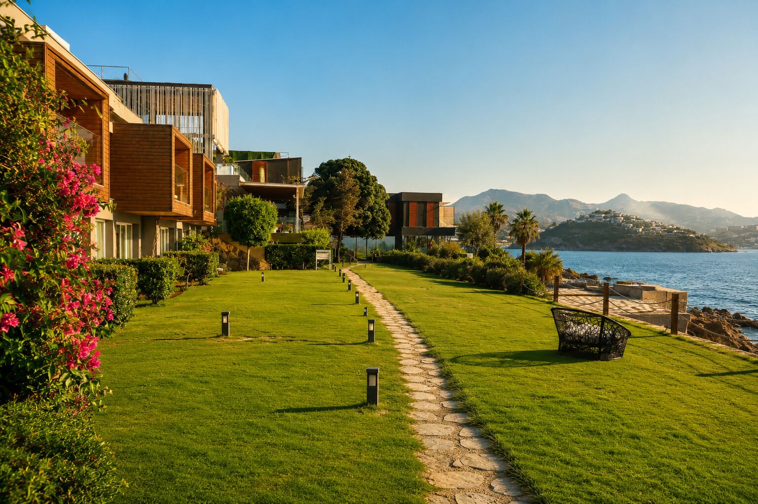 A coastal property with a paved pathway, modern buildings, and a view of the sea and mountains.