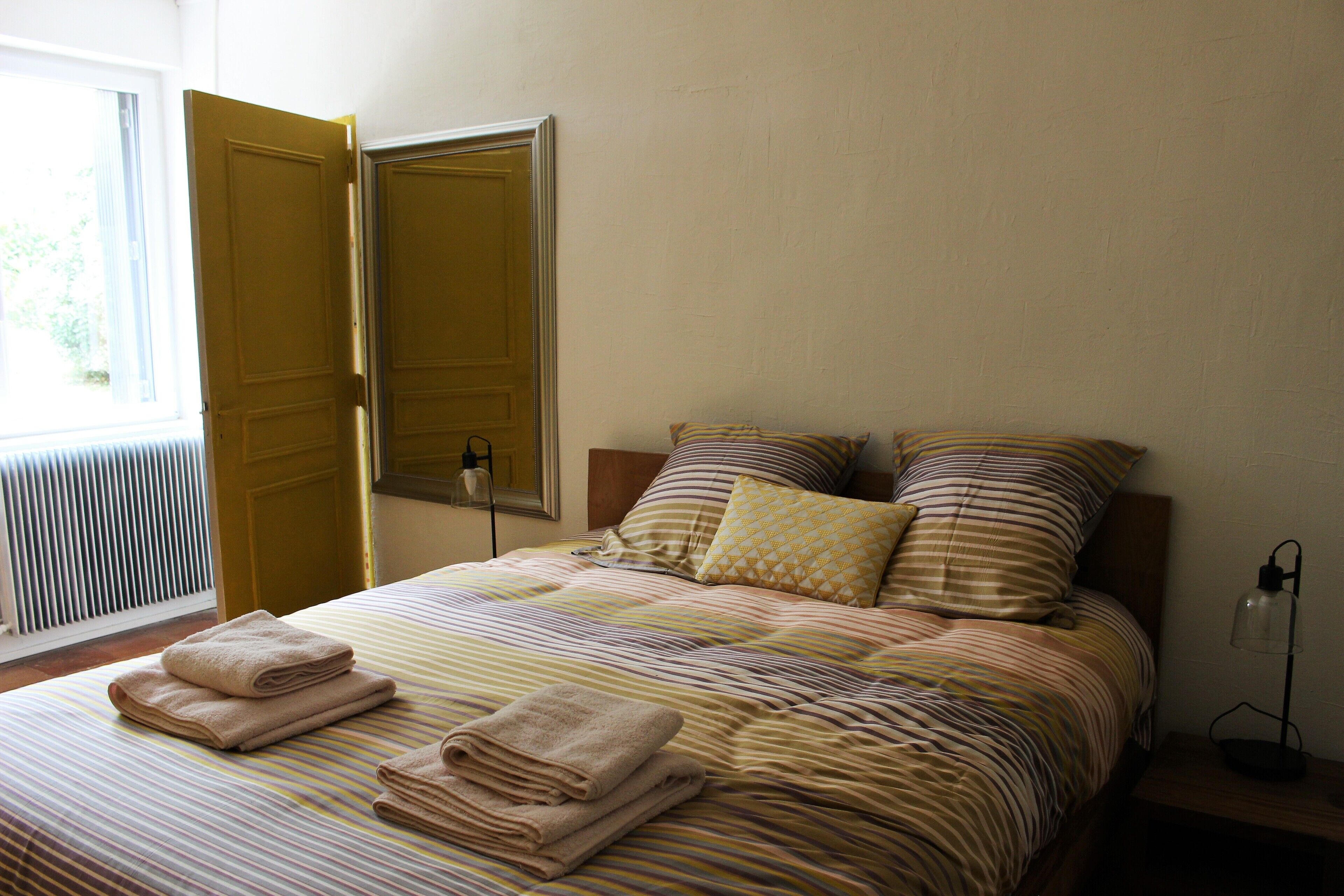 A neatly made bed with striped bedding and two pillows, a bedside lamp, and a window with blinds.