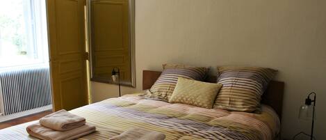 A neatly made bed with striped bedding and two pillows, a bedside lamp, and a window with blinds.
