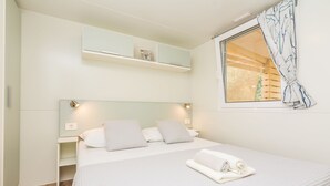 A bedroom with a bed, bedside tables, a window with curtains, and a shelf above the bed. - Mobile Home Pecten 04 (Pakostane)
