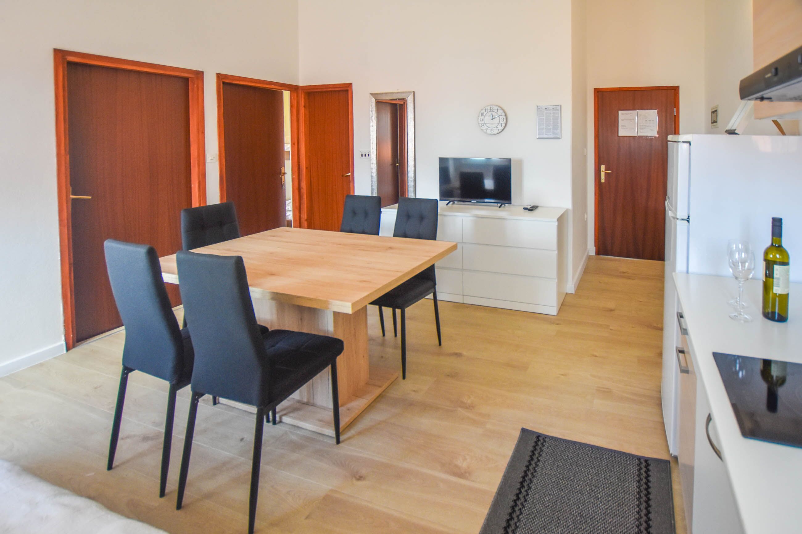 Comedor con mesa de madera y sillas negras, televisor, refrigerador y una botella de vino.