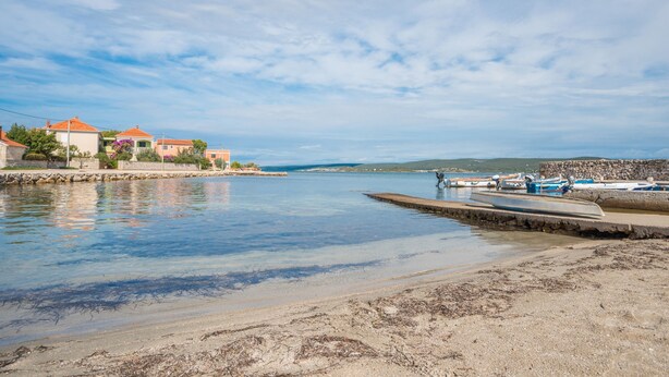 A coastal scene with a calm sea, a sandy beach, and a row of colorful buildings with red roofs. - Apartments Anchor (Pasman)