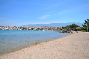 Eine Küstenansicht mit sandigem Strand, ruhigem Wasser, Booten und einer in der Ferne liegenden Stadt mit Bergen.