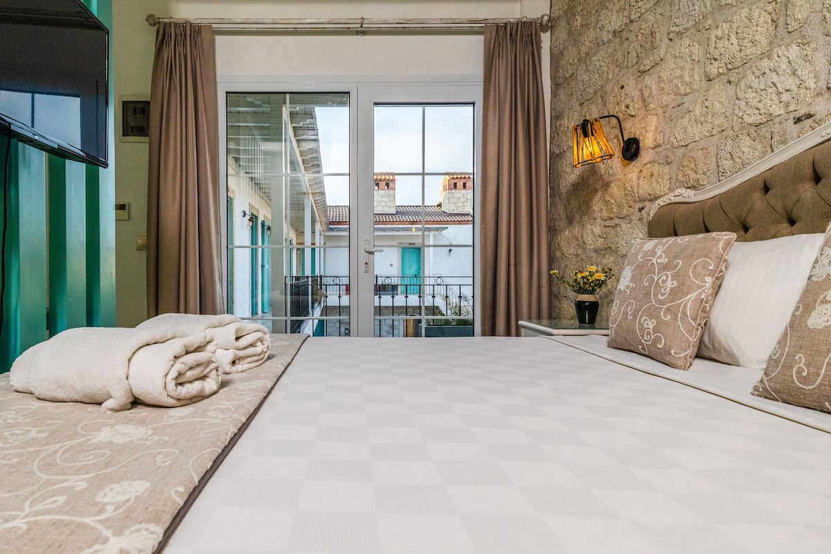A hotel room with a large bed, a television, a view of a building through the window, and a stone wall.
