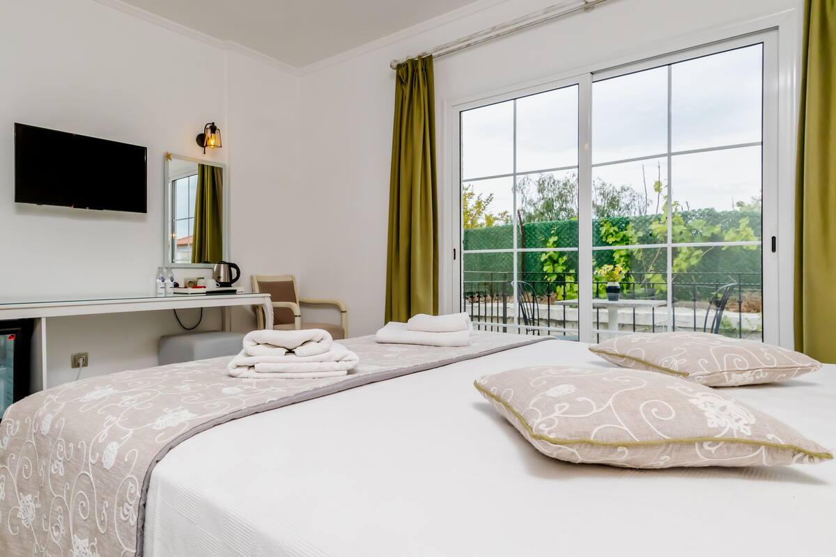 A hotel room with a bed, a desk, a television, and a view of greenery outside.