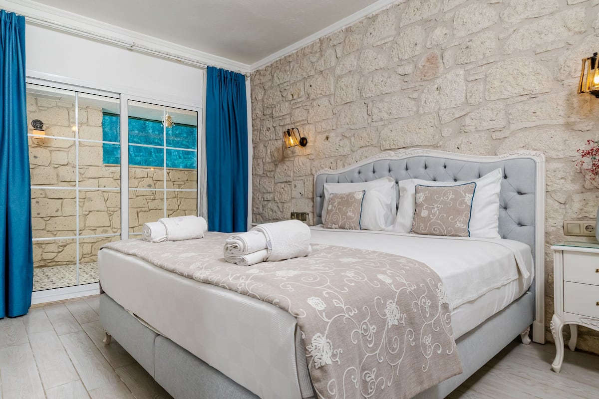 A bedroom with a stone wall, a large bed with a grey headboard, and a glass door leading to a pool.