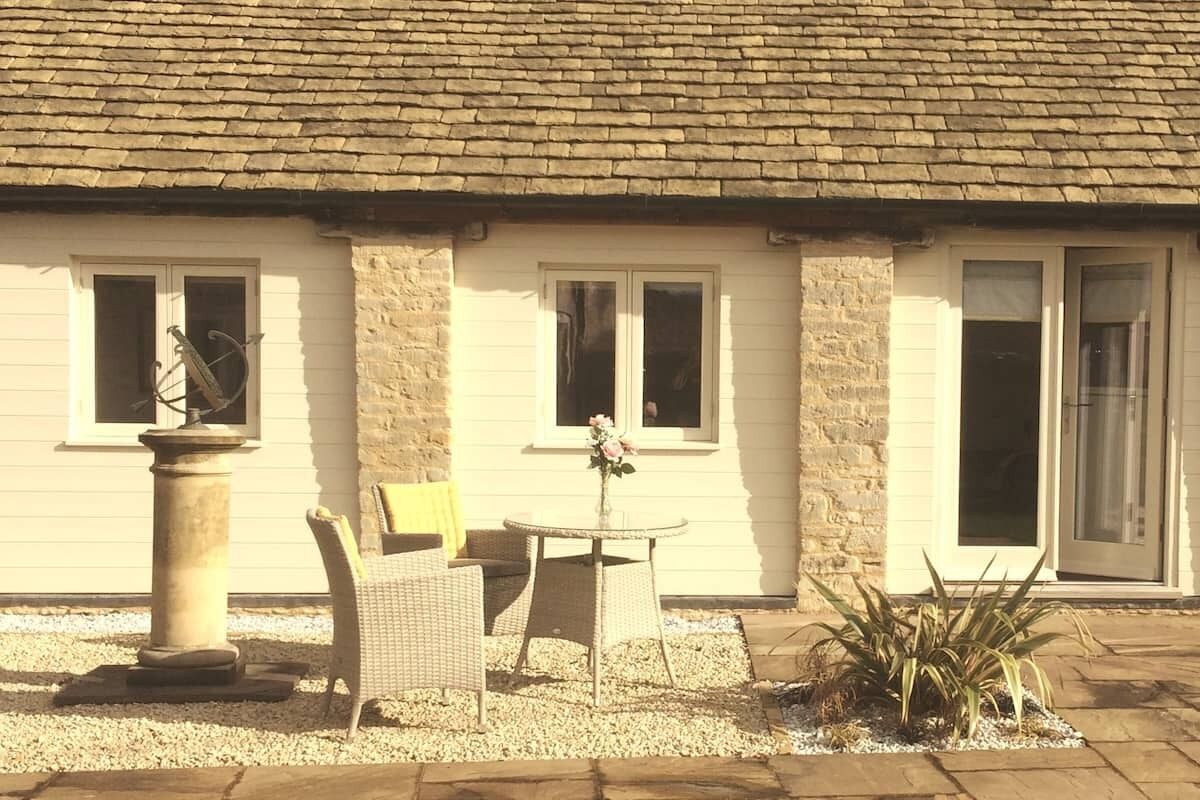 A building with a tiled roof, a stone wall, and a window with a flower arrangement. There is a wicker chair and a small table with a vase of flowers.