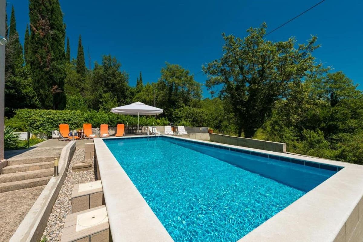 Ein rechteckiger Swimmingpool mit klarem, blauem Wasser, umgeben von einer Steindeck und Treppen. Mehrere Liegestühle und ein weißer Sonnenschirm sind vorhanden.