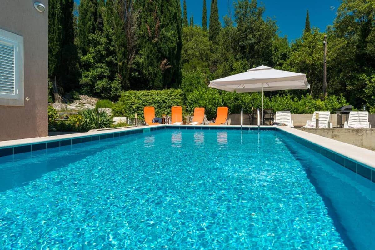 Ein rechteckiger Swimmingpool mit klarem, blauem Wasser, umgeben von orangefarbenen Liegestühlen und einem weißen Sonnenschirm.