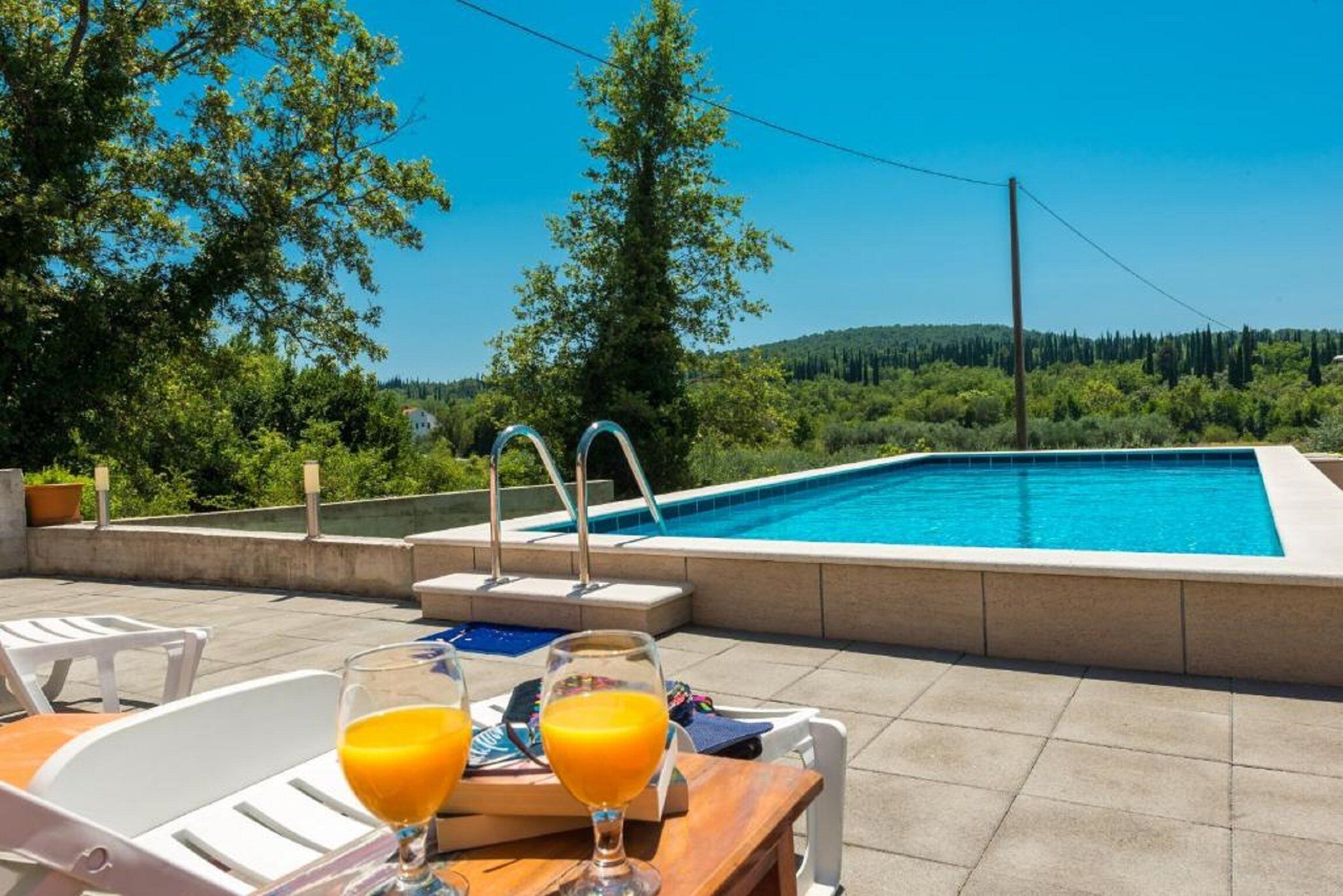 A swimming pool with a view of trees and hills, two glasses of orange juice on a table, and a clear blue sky.