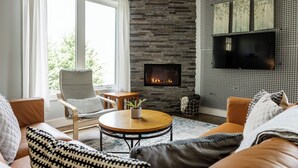 A living room with a stone fireplace, a flat-screen TV mounted on a grid-patterned wall, and a large window with curtains. - Eco- Friendly house near UNCA and downtown AVL, Mountain Views  (Asheville)