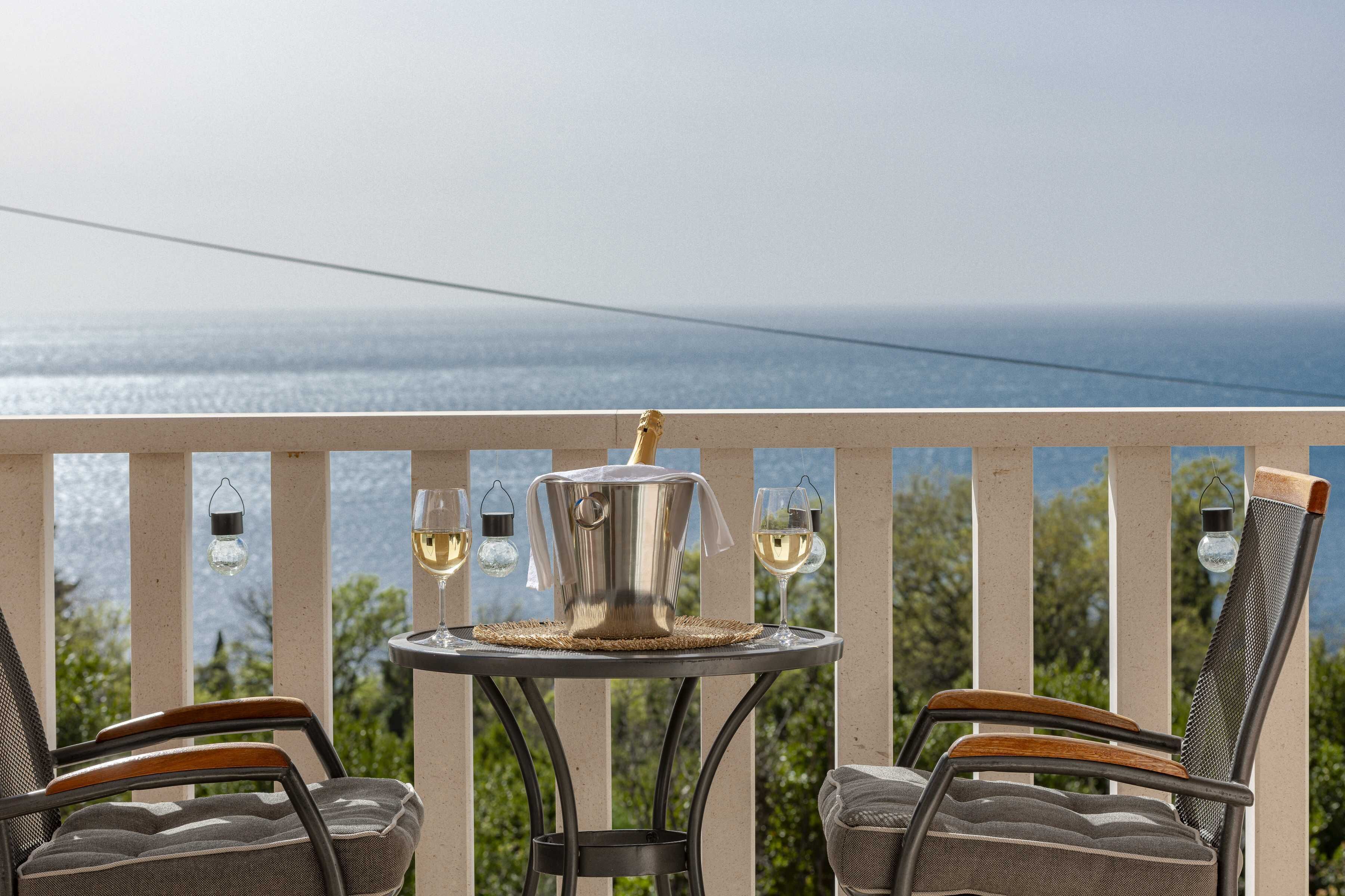 A balcony with a table set for two, featuring a metal pot and two glasses of white wine, overlooking a body of water.