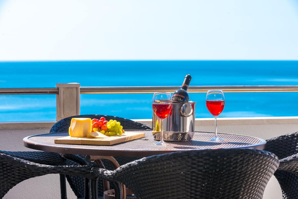 A table with a bottle of wine, two glasses, a cheese platter, and grapes, set against a view of the ocean.