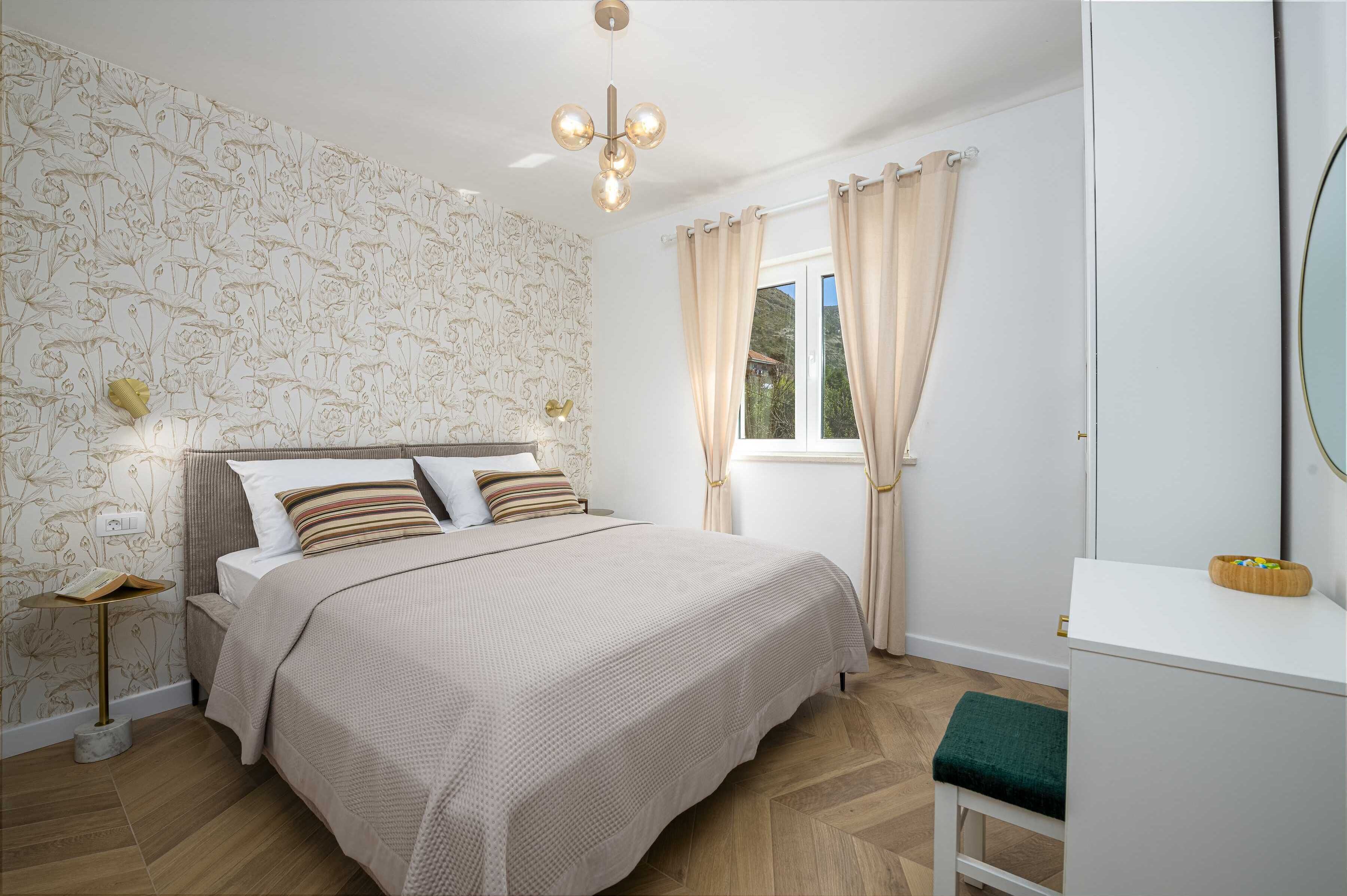 A bedroom with a large bed, a bedside table with a lamp, a window with curtains, and a mirror on the wall.