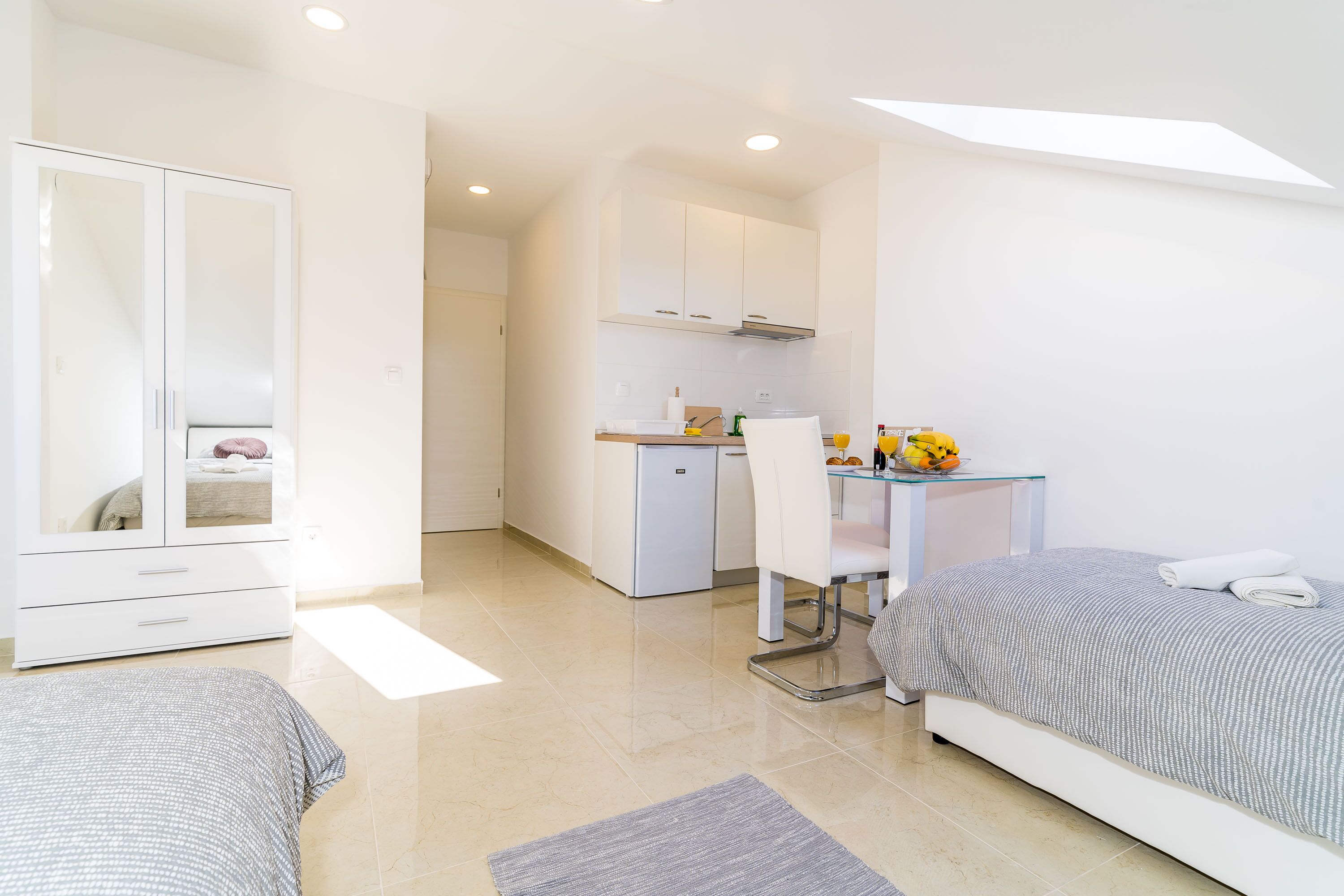 Quarto moderno e bem iluminado com cama, mesa de cabeceira e guarda-roupa. Uma pequena cozinha compacta é visível ao fundo.
