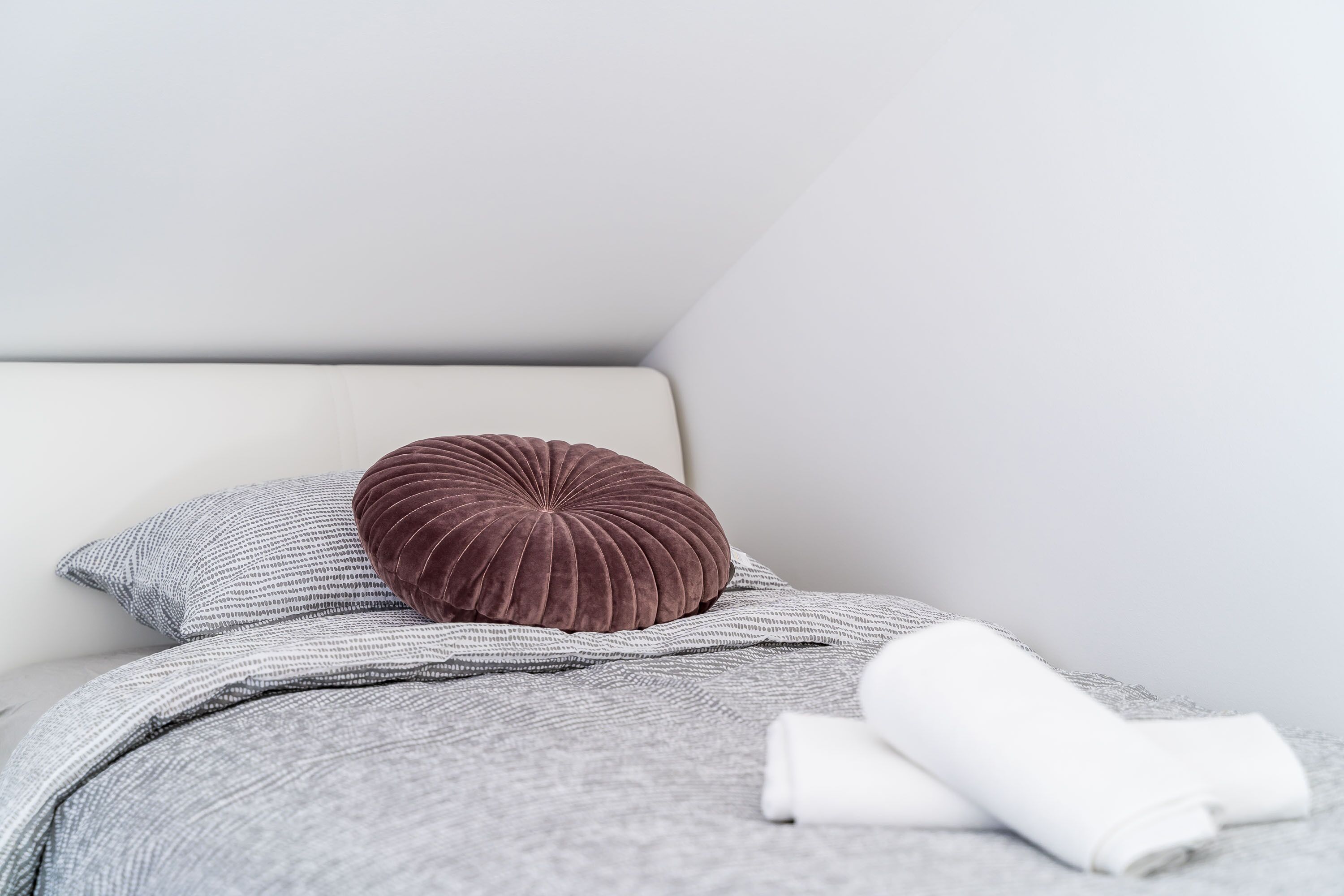 A neatly made bed with a grey blanket, a brown pillow, and a rolled white towel.