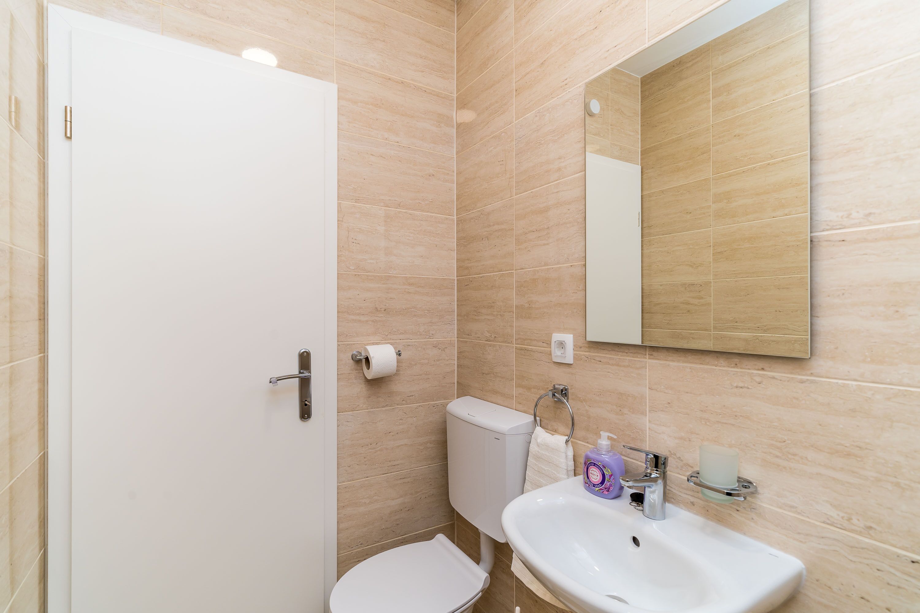 A bathroom with beige tiles, a white toilet, a sink with a mirror above it, and a toilet paper holder.