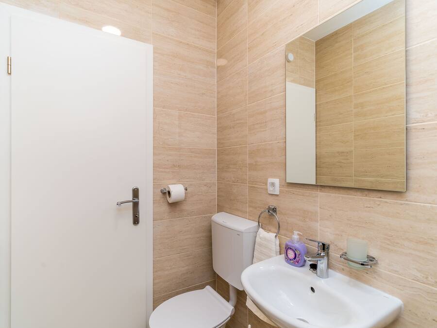 Casa de banho com azulejos bege, sanita branca, lavatório com espelho em cima e suporte para papel higiénico.