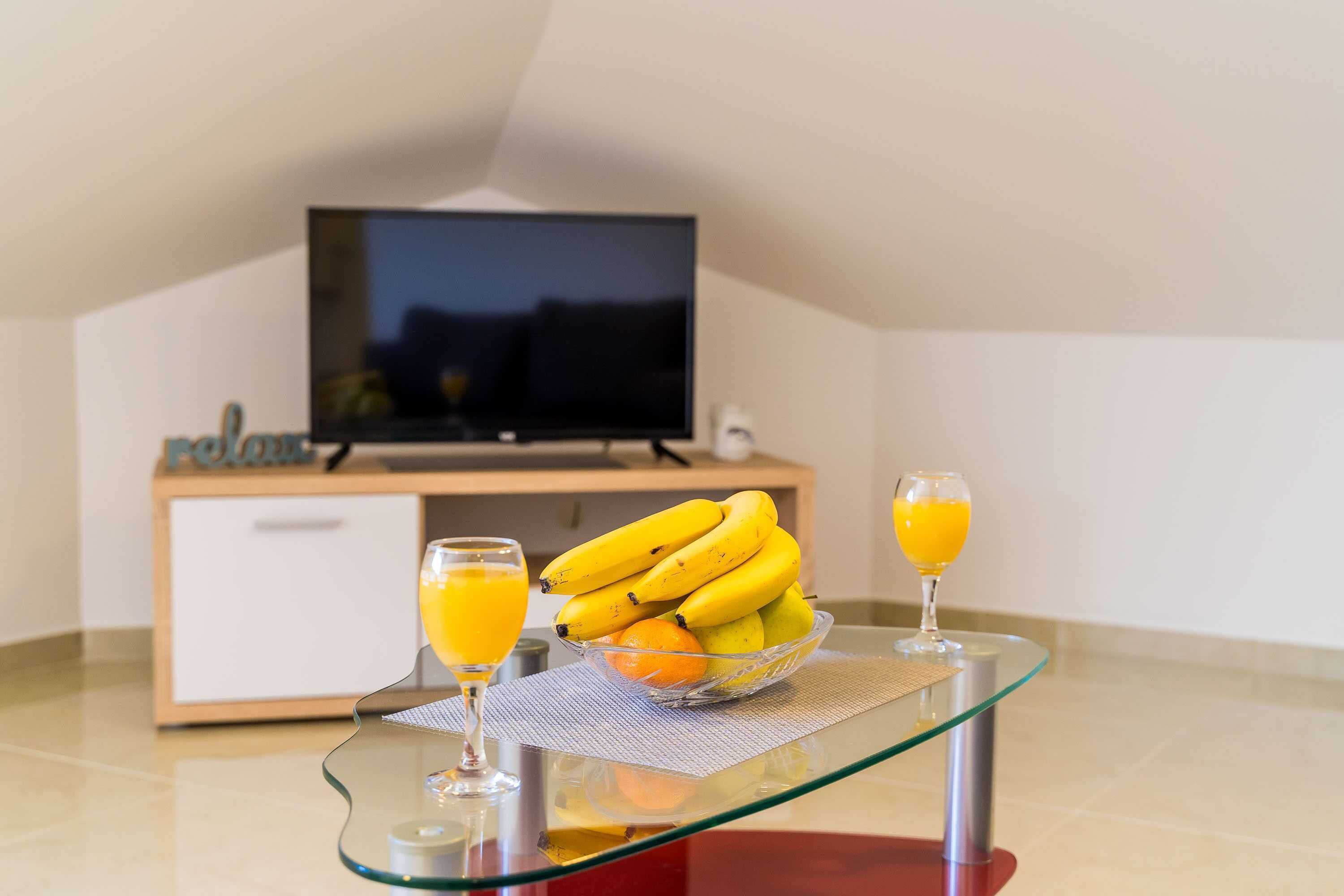Una mesa de vidrio con un frutero y dos vasos de jugo de naranja, un televisor y un aparador de madera al fondo.