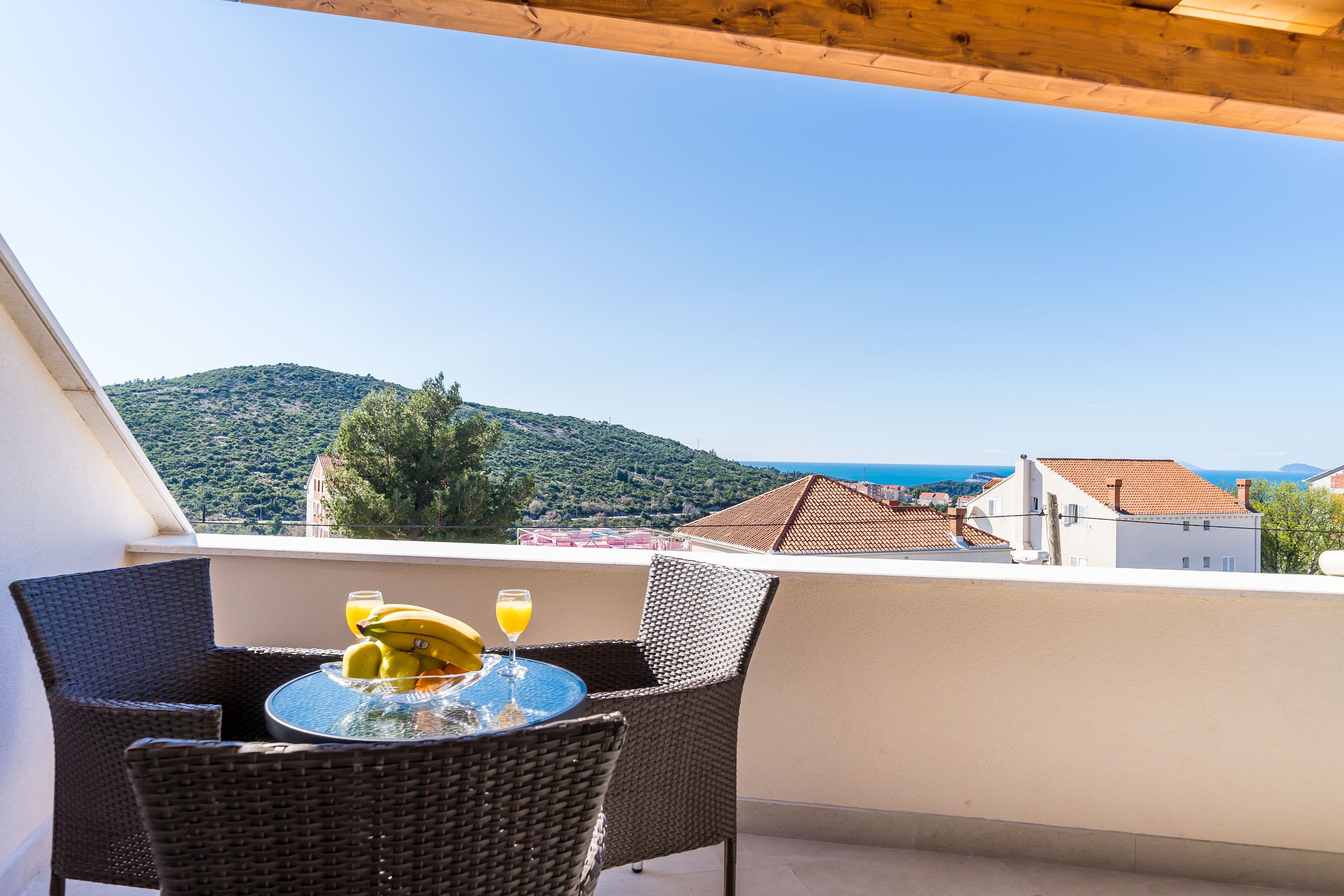 A balcony with wicker chairs and a table, overlooking a hillside and buildings.
