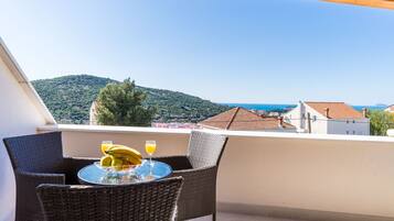 A balcony with wicker chairs and a table, overlooking a hillside and buildings.