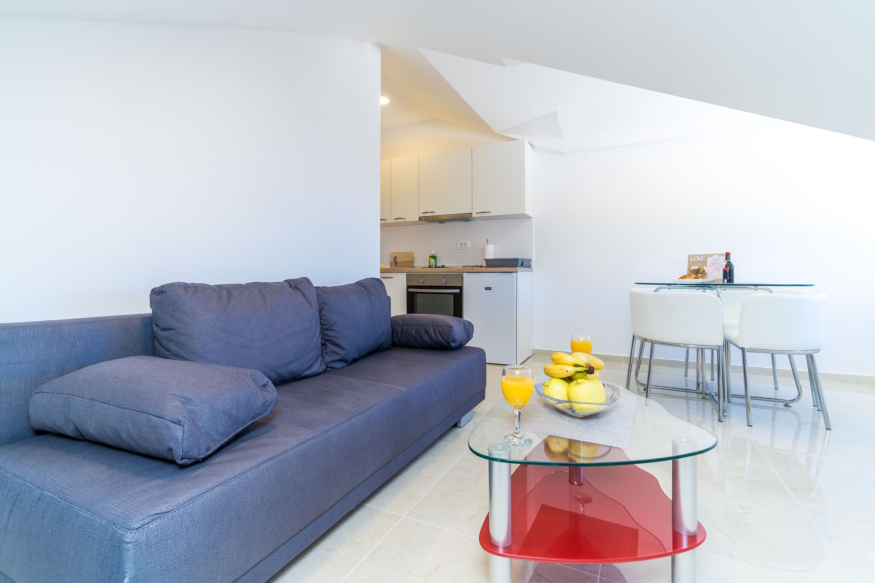 Una sala de estar moderna con un sofá gris, una mesa de centro de vidrio con base roja, y una cocina al fondo.