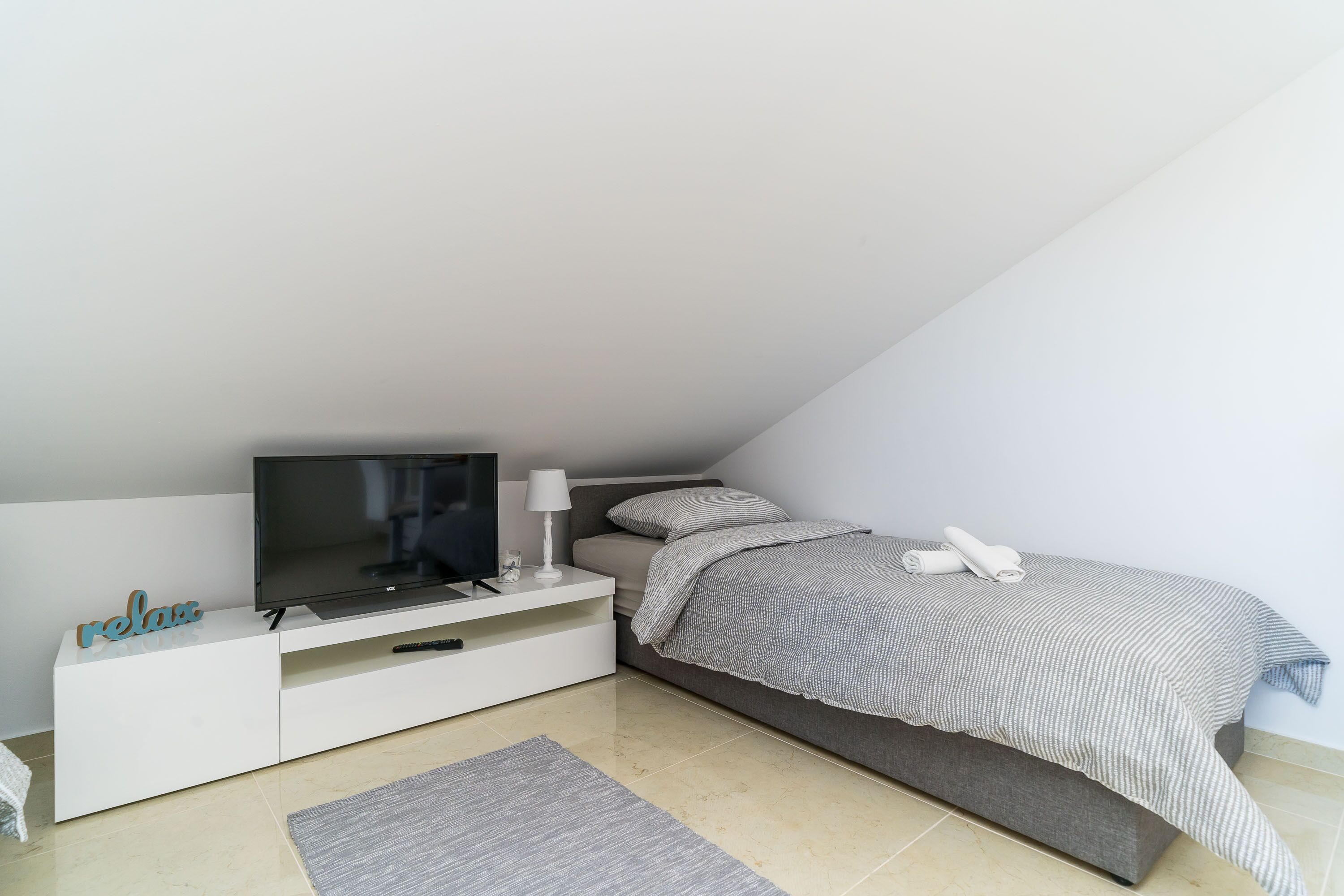 Un dormitorio moderno con una cama, un televisor sobre un soporte blanco, una mesita con una lámpara y una alfombra en el suelo.