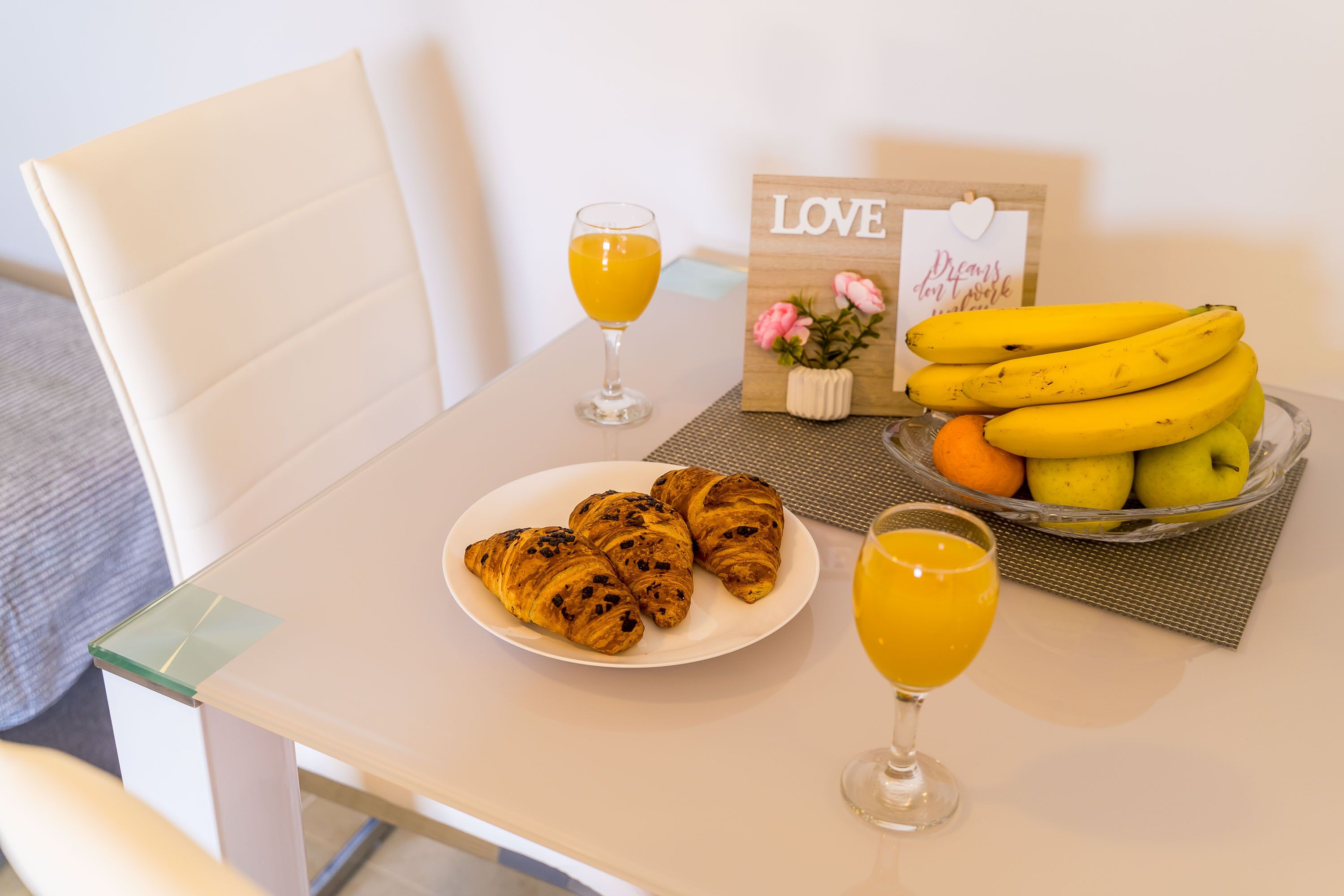 Uma mesa com croissants, um copo de sumo de laranja, um copo de sumo de laranja, uma taça de bananas e um prato de croissants.