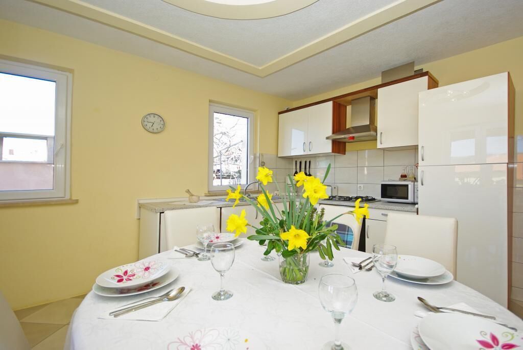 A dining area with a table set for four, a vase of yellow flowers, and a kitchen in the background.
