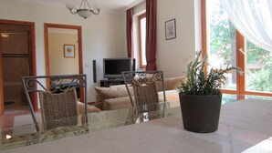 Ein Essbereich mit einem Glastisch, Korbstühlen, einer Topfpflanze, einer Couch und einem Fernseher im Hintergrund.