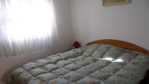 Een bed met een bloemenbedekking in een kamer met een effen muur, een raam met dunne gordijnen en een klein nachtkastje met een lamp.