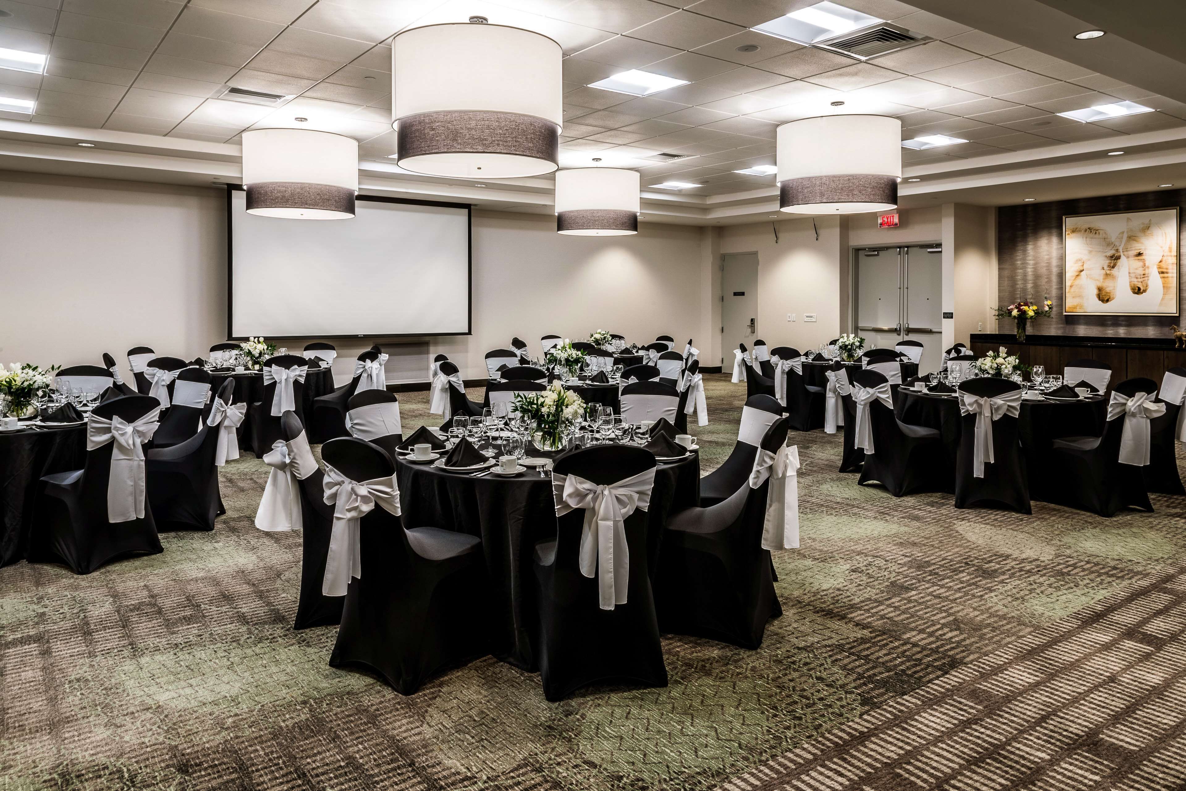 A conference room with multiple tables set for a formal event, featuring black chairs with white sashes, large hanging lamps, and a projector screen.