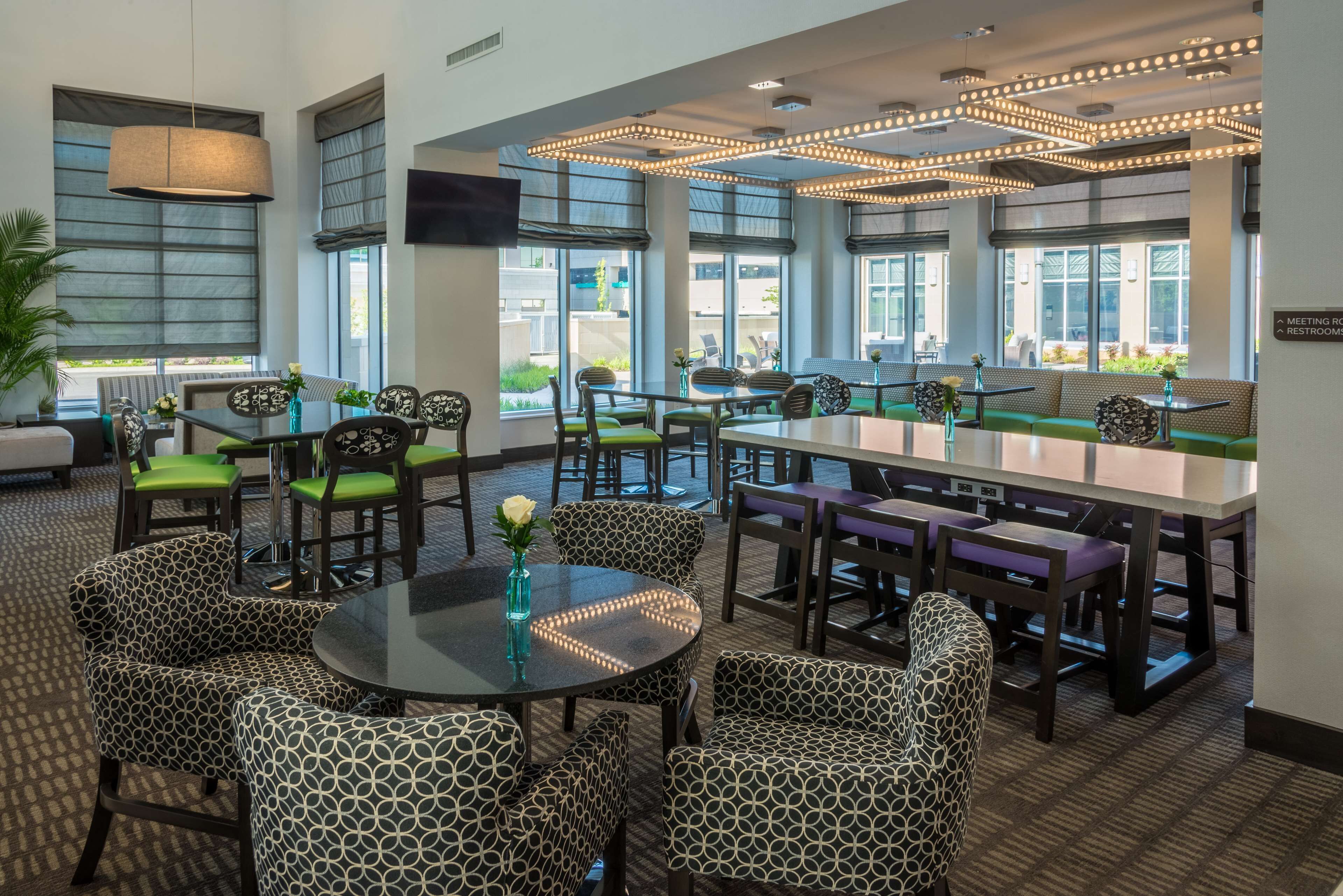A modern restaurant with patterned chairs and tables, large windows, and a ceiling light fixture.