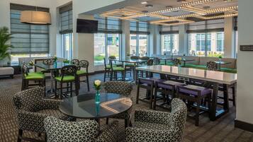 A modern restaurant with patterned chairs and tables, large windows, and a ceiling light fixture.