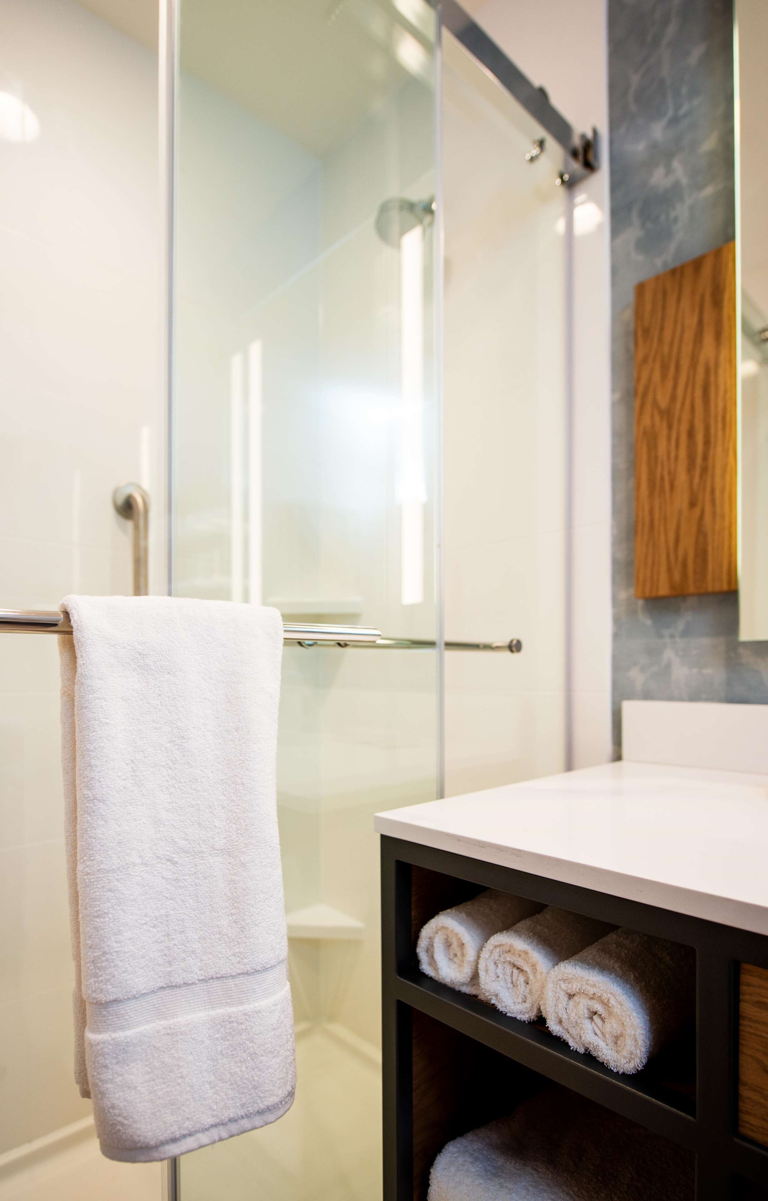 A modern bathroom with a glass shower enclosure, a white towel hanging on a metal bar, and neatly rolled towels on a shelf.