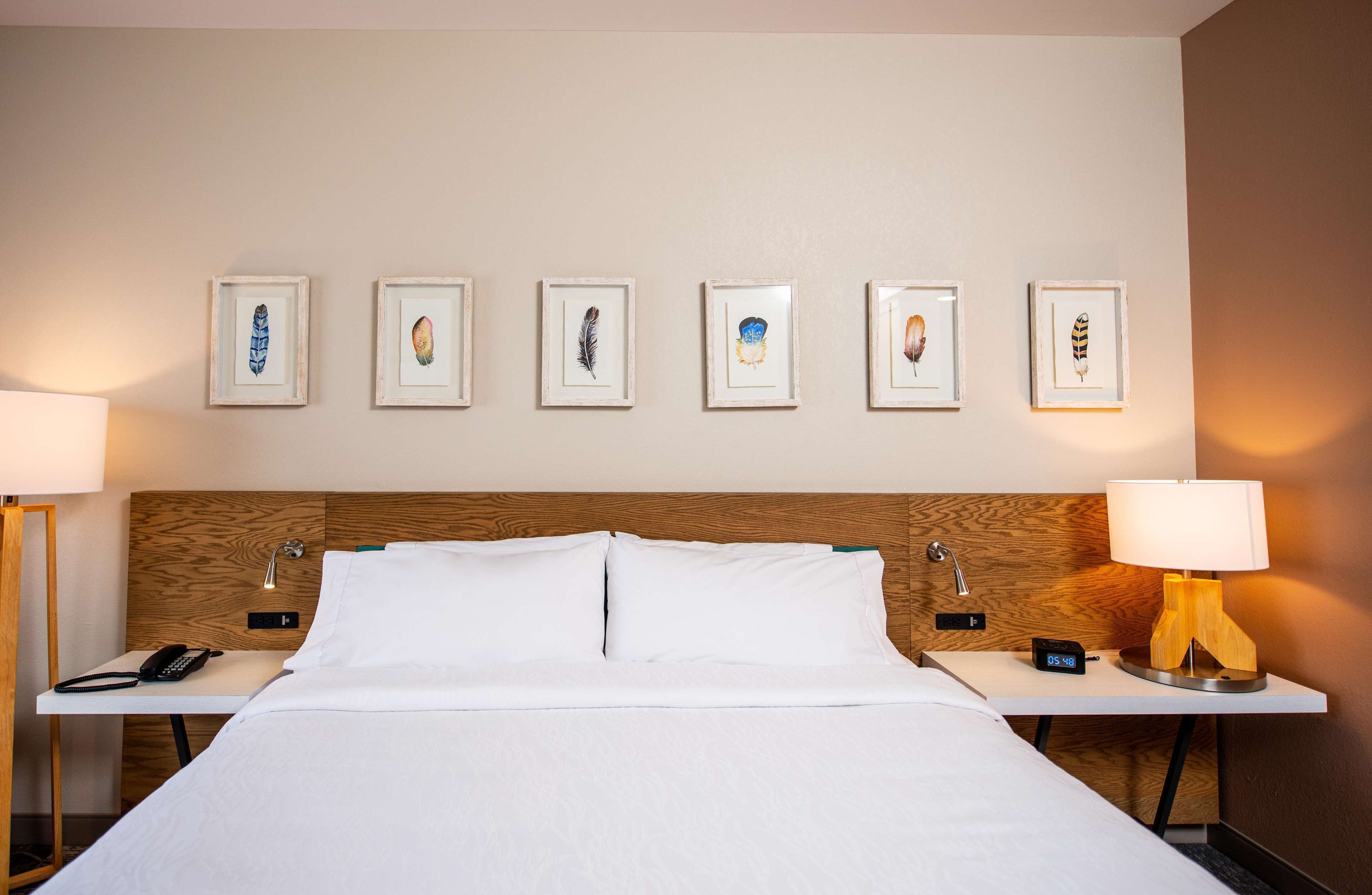 A hotel room with a neatly made bed, two bedside tables with lamps, a nightstand, and a wall with six framed artworks.