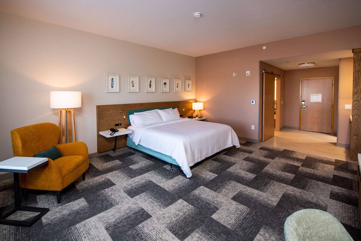 A hotel room with a bed, a desk, a chair, a nightstand, a lamp, and a door leading to another room.