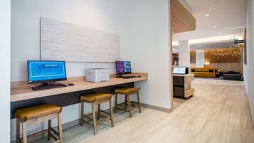 Eine moderne Hotellobby mit einer Rezeption, einem Computerarbeitsplatz mit zwei Monitoren und einer Sitzecke mit Holzstühlen.