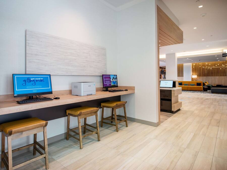 Eine moderne Hotellobby mit einer Rezeption, einem Computerarbeitsplatz mit zwei Monitoren und einer Sitzecke mit Holzstühlen.