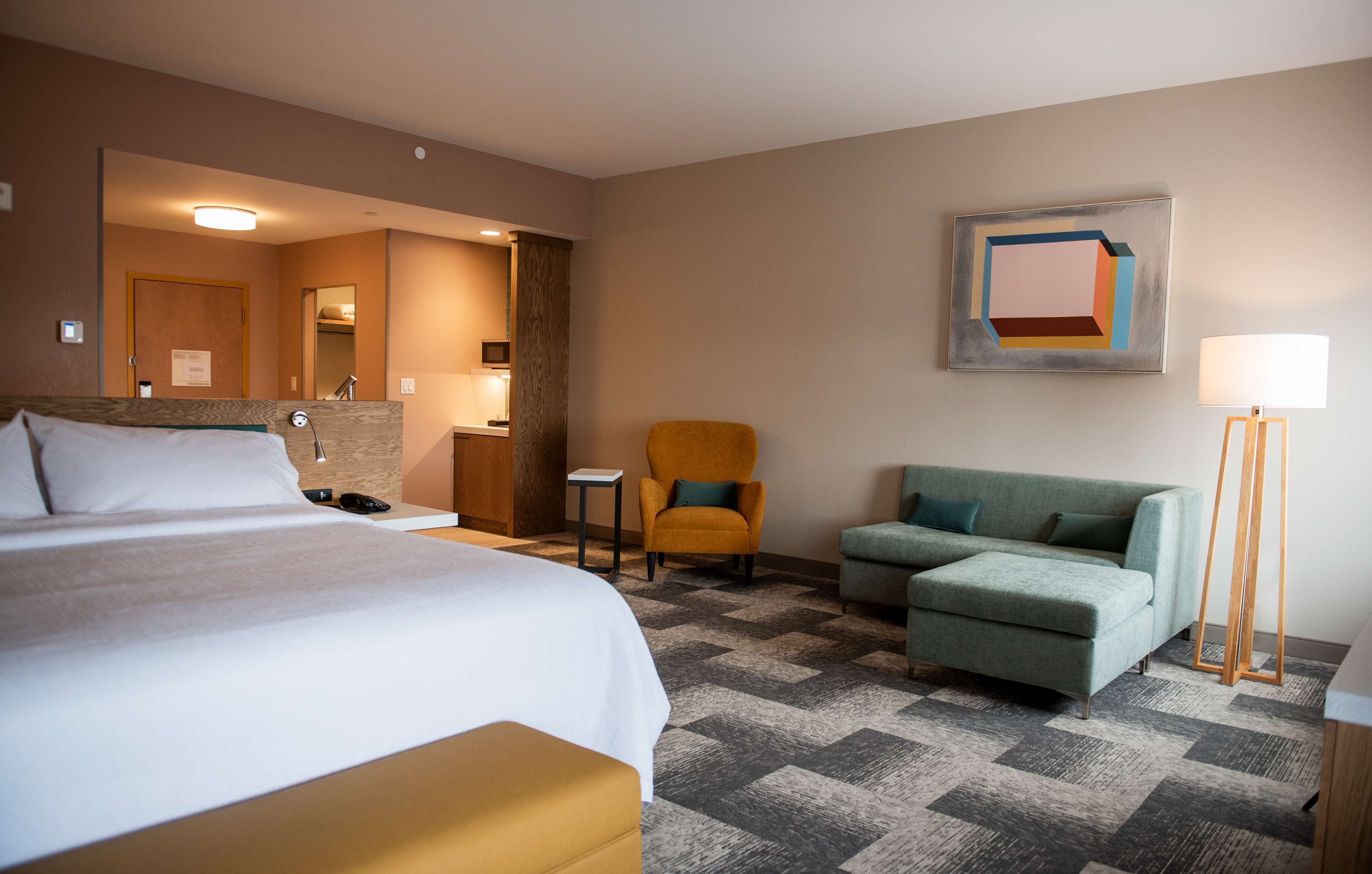 Habitación de hotel con cama, sofá, sillón, escritorio, lámpara y un cuadro en la pared.