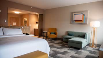 Habitación de hotel con cama, sofá, sillón, escritorio, lámpara y un cuadro en la pared.