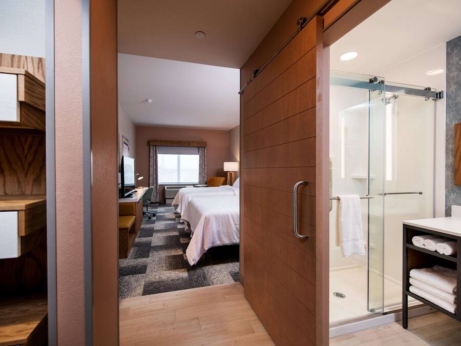 Habitació d'hotel moderna amb un armari encastat, una dutxa amb porta de vidre, un llit amb edredó blanc i un escriptori amb cadira.