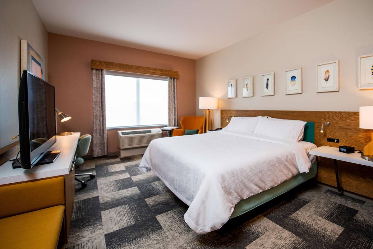 A hotel room with a large bed, a desk with a computer, a chair, a TV, and a window with curtains.