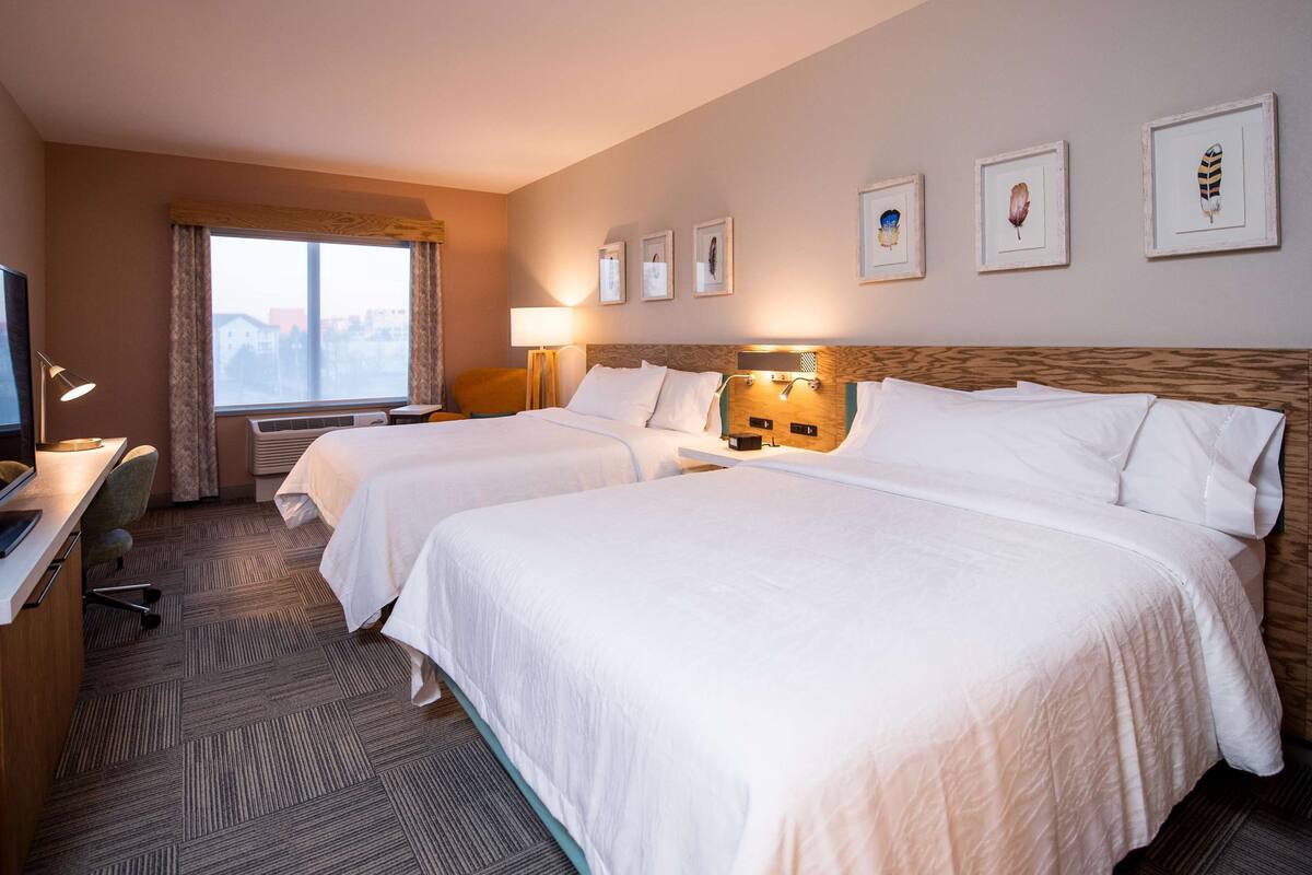 A hotel room with two beds, a desk with a computer, a TV, and framed artwork on the wall.