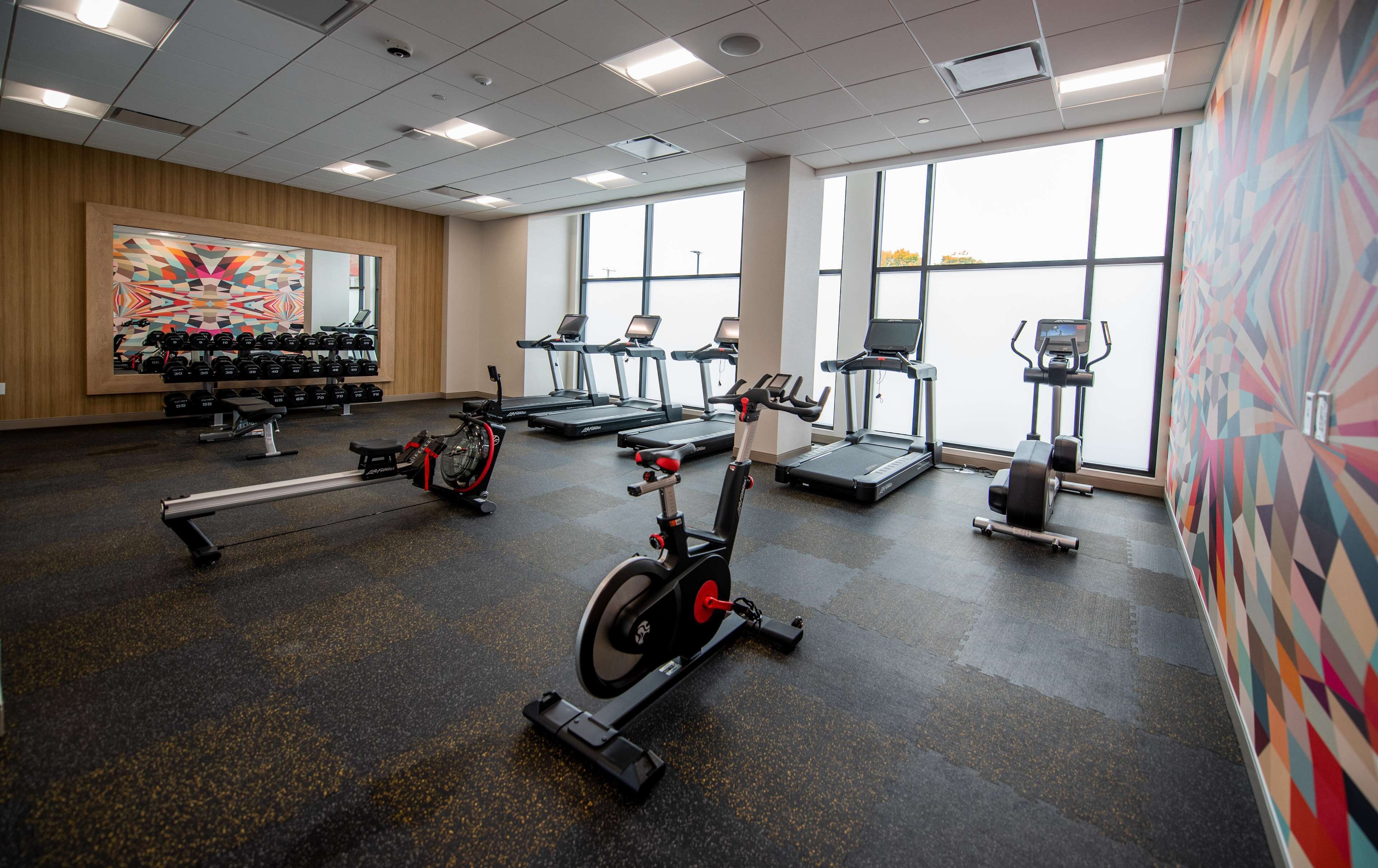 Un gimnasio con cintas de correr, bicicletas estáticas y una máquina de remo. Hay mancuernas en estanterías y un amplio ventanal con vistas al exterior.