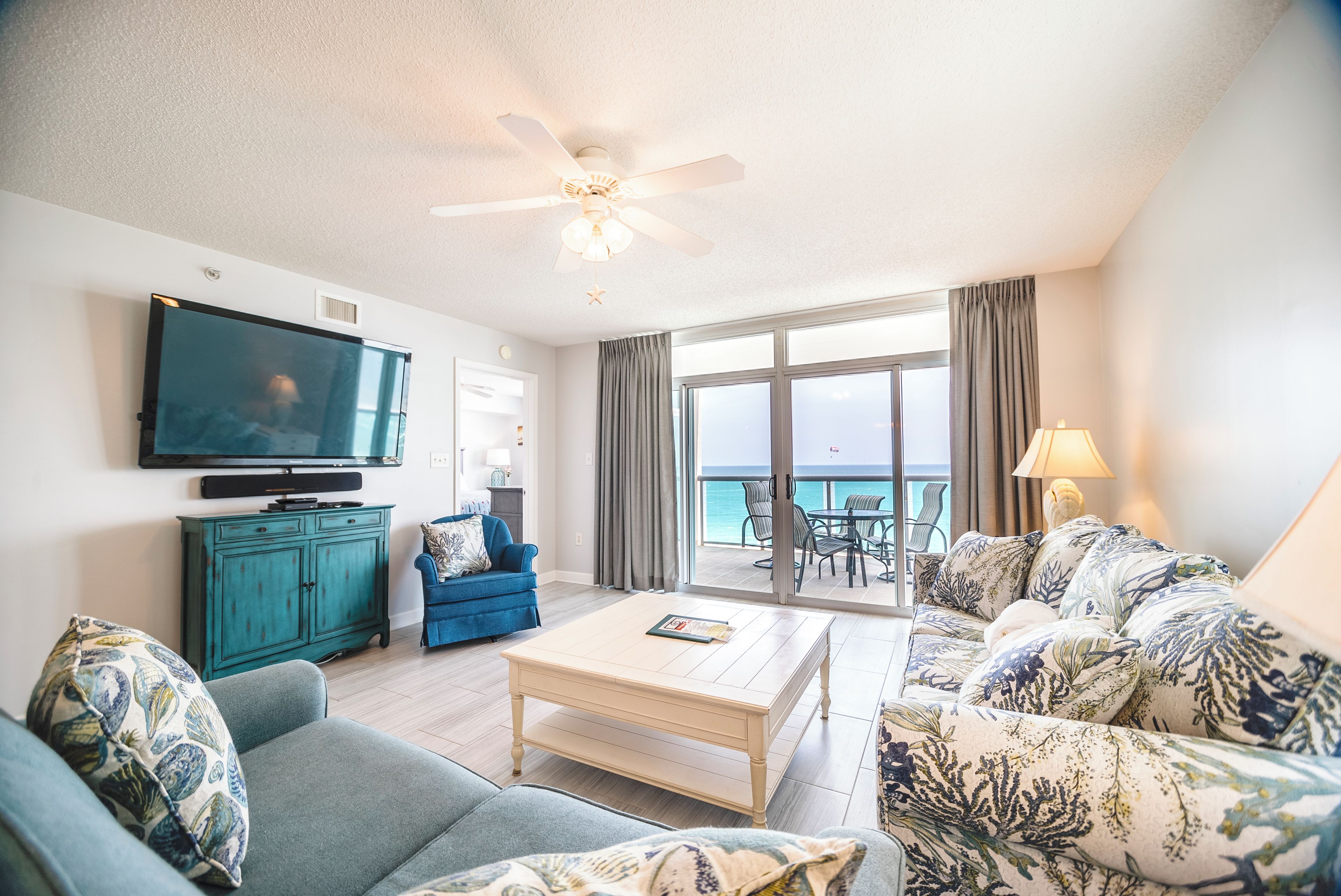 A living room with a flat-screen TV, a ceiling fan, a sofa, a coffee table, and a view of the ocean through large windows.